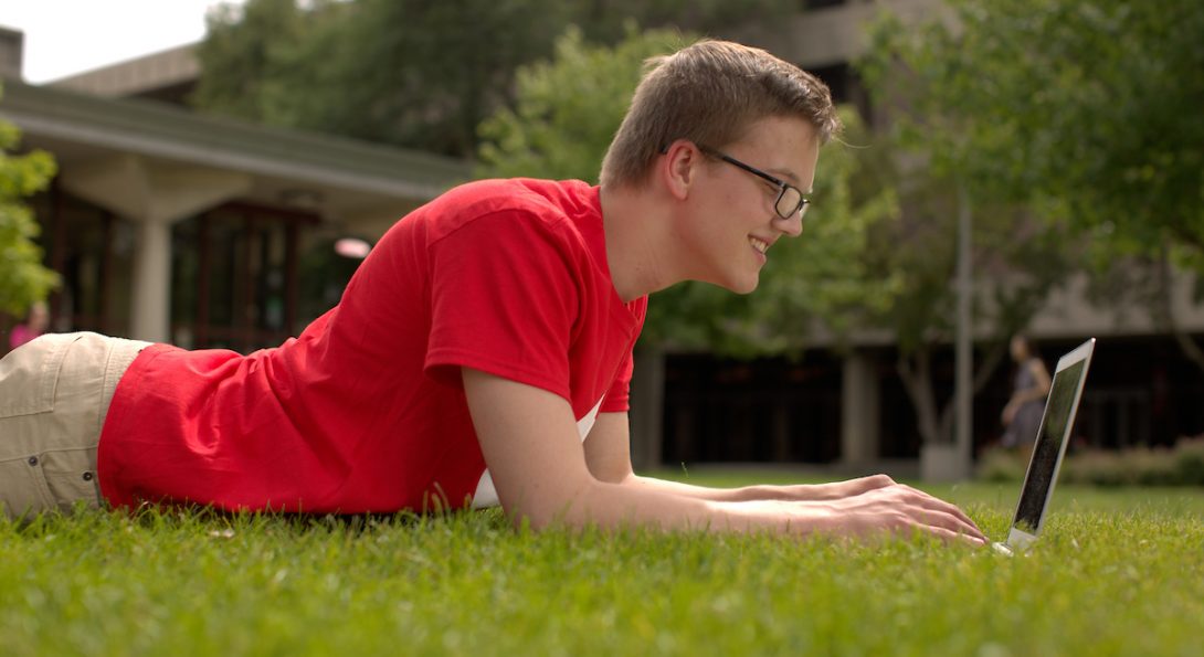 Male student laying on lawn with a laptop