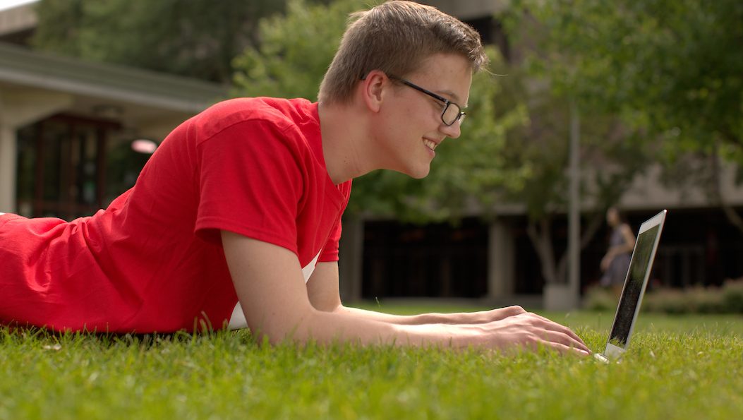 Male student laying on lawn with a laptop