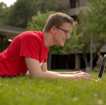 Male student laying on lawn with a laptop
                  