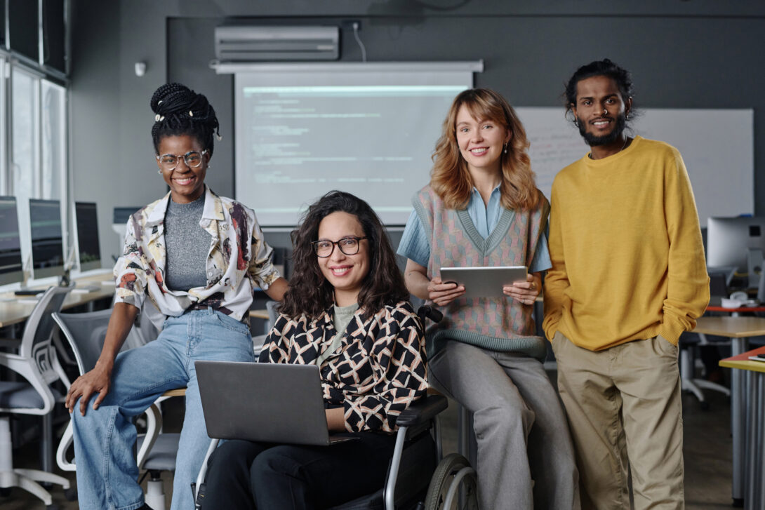 Four students representing diverse ethnicities in a classroom, two holding technology (laptop and tablet), one in a wheelchair.
